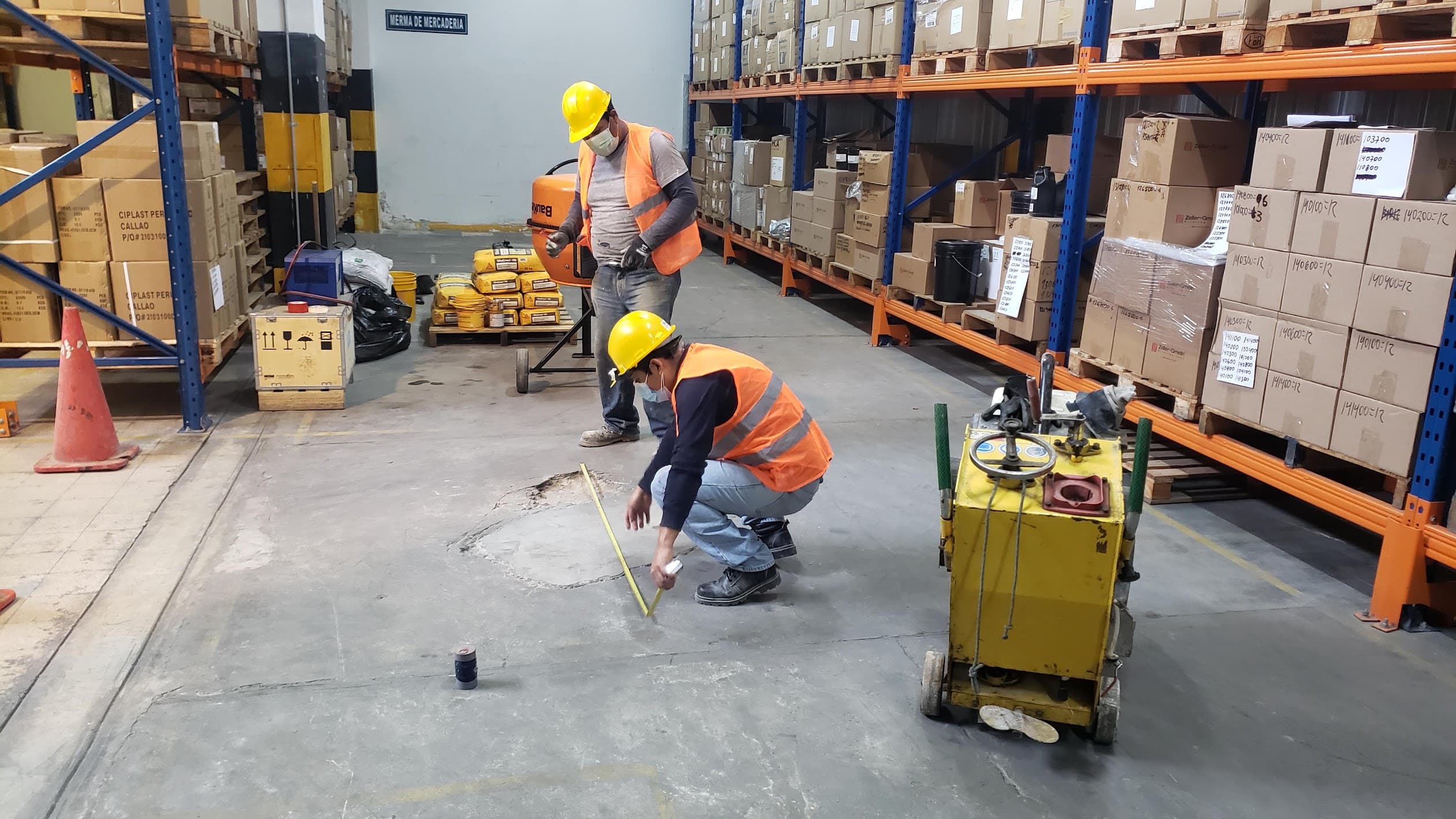 Dos técnicos con casco y chaleco evalúan y miden un daño en el piso de un almacén industrial